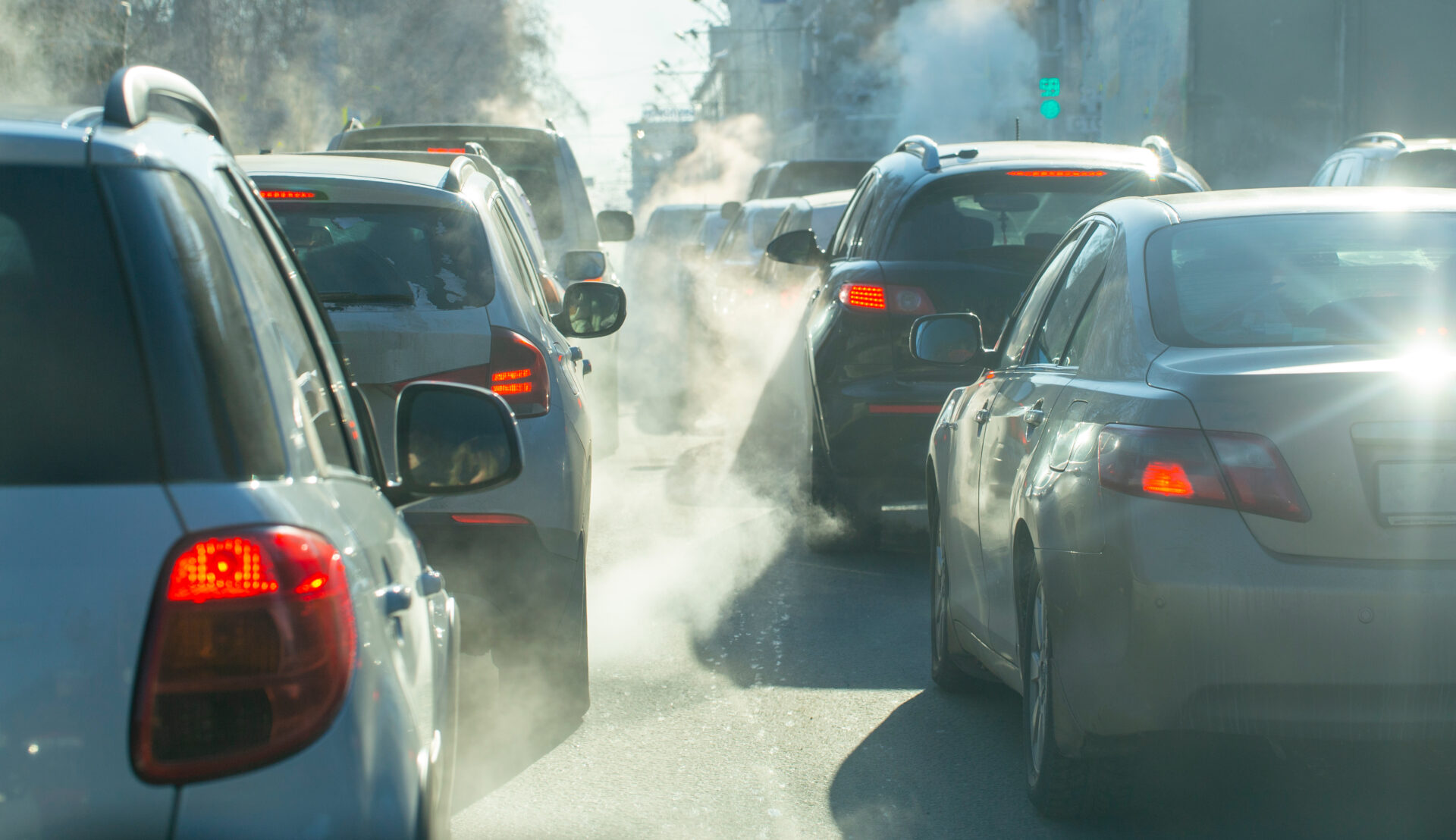 Exhaust from cars fills the frame on a cold winter morning. Silver and white cars are lined up in traffic, idling.