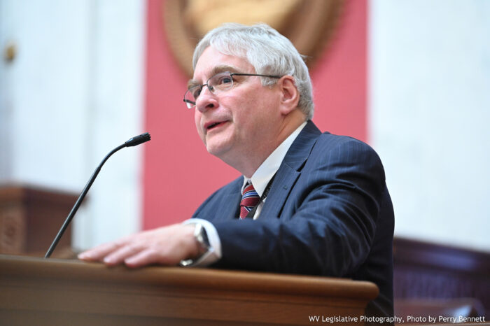 An older man with white, salt and pepper hair, stands at a podium and speaks into a microphone. He is speaking from the West Virginia House of Delegates floor.