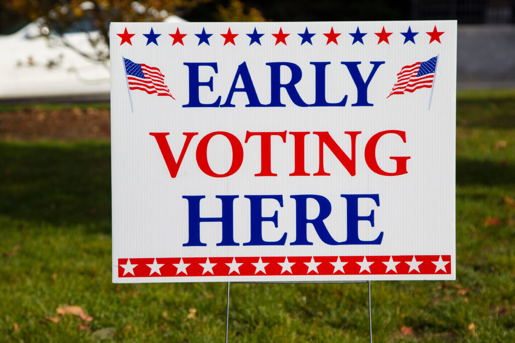 A close up photo of a sign that reads, "Early Voting Here." Around the word "early" are two American flags. The colors on the sign feature red, white and blue.