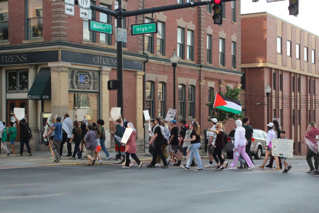A group holding signs walks in front of a building labeled "Citizens" and under green street signs that read "Walnut" and "High". A woman in the group holds up a pole with the Palestinian flag, a red triangle on the left with black, white and green stripes. A child holds a sign to the camera that reads "Free Gaza"
