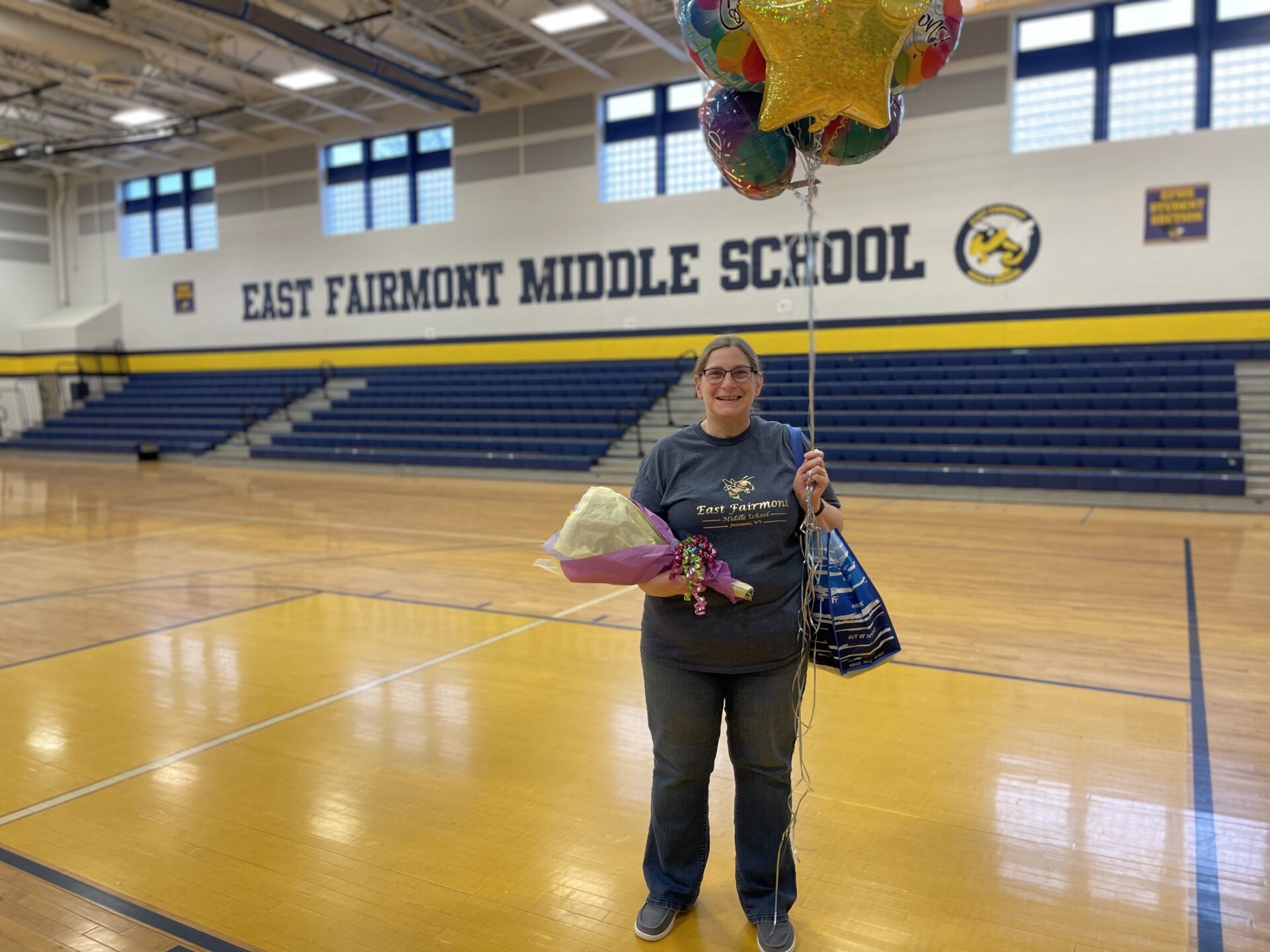 An older woman stands in the center of an empty school gymnasium. She holds a bouquet in one hand and a tote bag in the other. She is smiling for the camera, has her salt and pepper hair pulled back into a ponytail, and wears glasses.