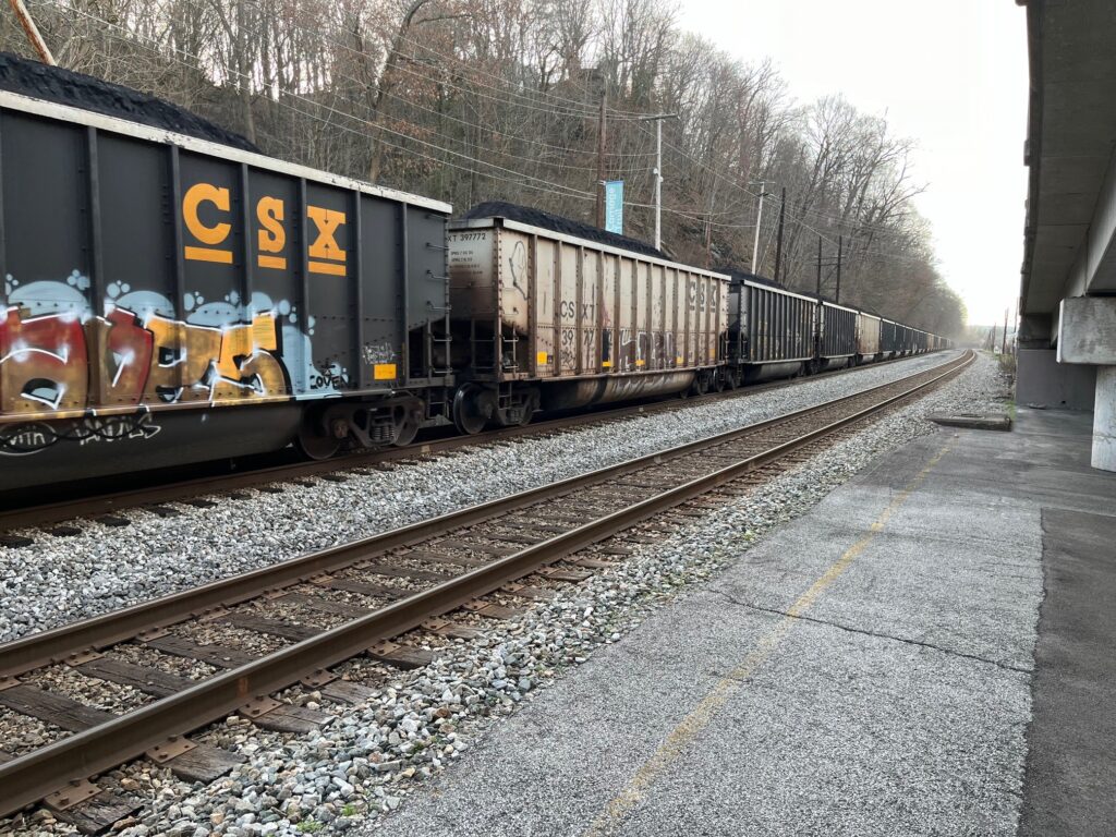 A train of black and gray railcars carrying coal passes through an Amtrak station on an overcast day.