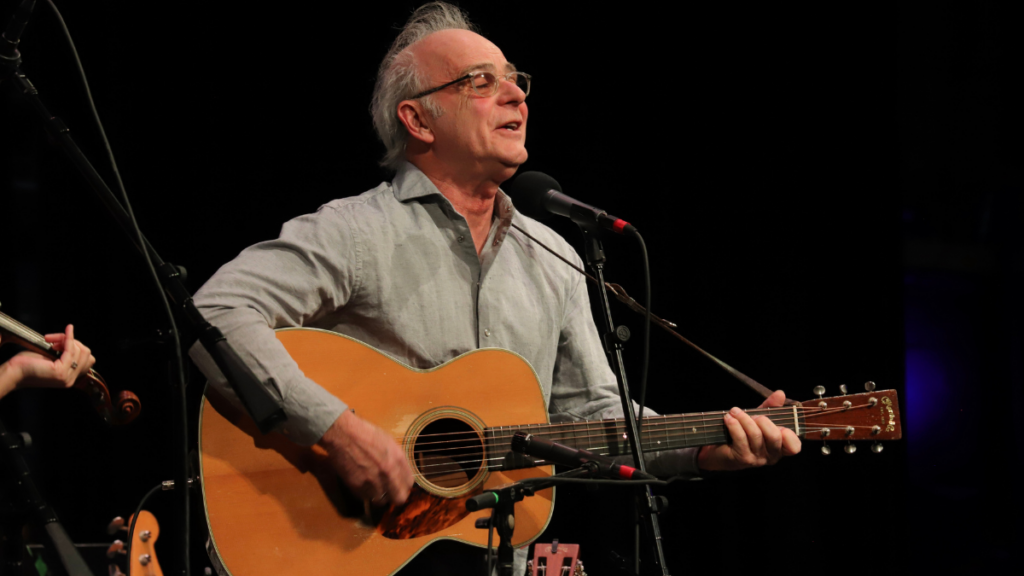 An adult man with graying hair and glasses. He holds an acoustic guitar.