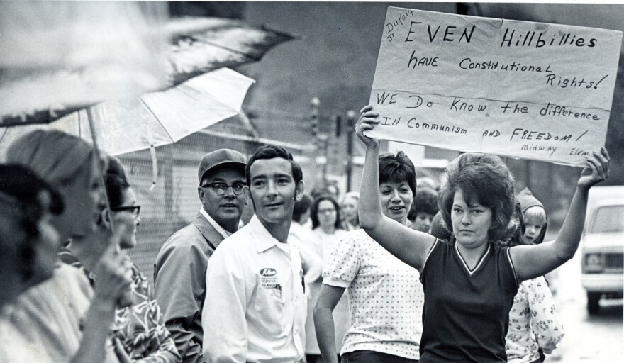 Protesters stand along a road, dressed in clothes from the 1970s. A woman holds up a sign that reads "Even hillbillies have constitutional rights! We do know the difference in communism and freedom!"