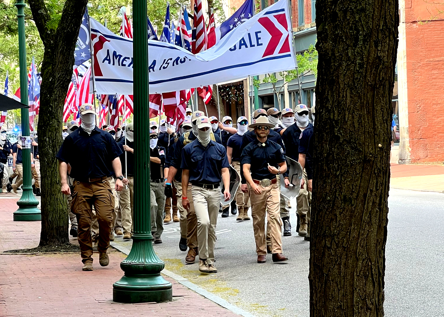 Several white men are shown dressed in navy shirts and khakis. The men wear ball caps and white face masks to hide their faces. They hold upside down American flags and march down a street during the daytime.