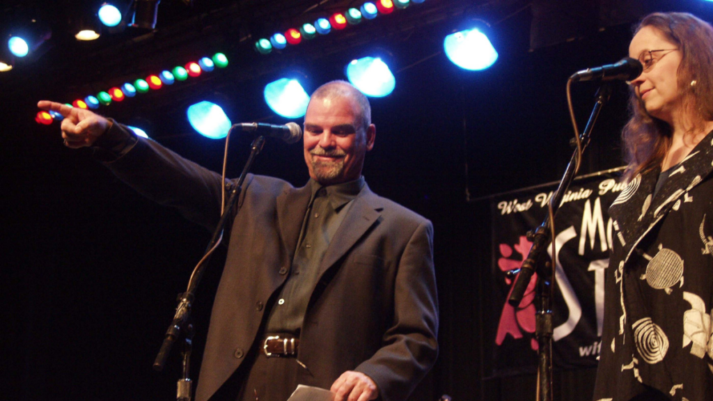 A man stands on a stage, looking down, while pointing his right index finger out toward the audience and smiling. Next to him is a woman in front of a microphone.