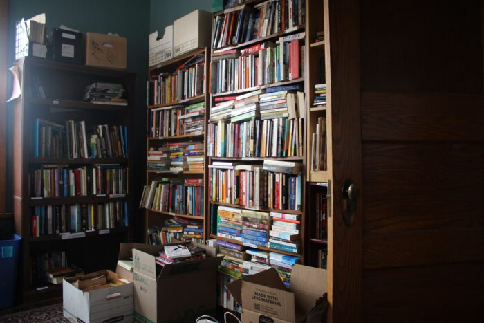 Cardboard boxes sit on an oriental carpet on the floor in front of floor to ceiling shelves packed with multicolored paperback books. Light streams in from the left of frame.