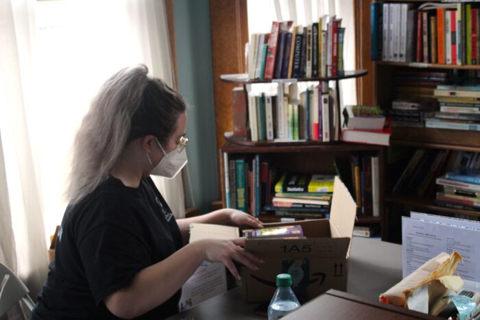 A woman with a high ponytail wearing glasses, a white N95 mask and a black shirt sits at a table while opening a cardboard Amazon shipping box filled with books. She sits in front of a window draped with sheer white curtains with daylight streaming through. To the right in the background are shelves covered in books.