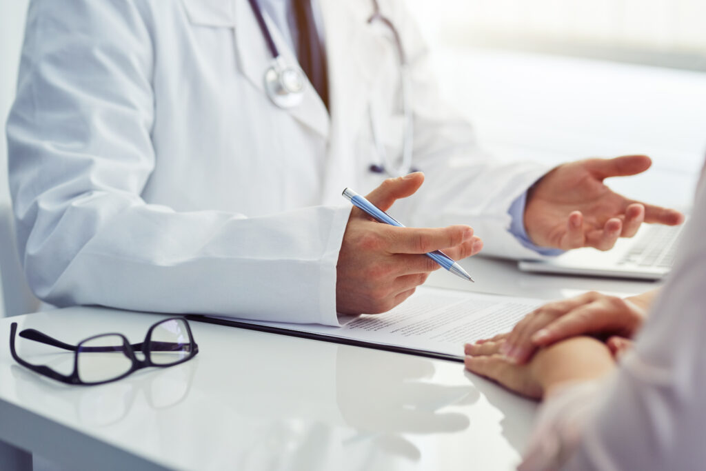 A white male doctor is seen from the neck down, speaking with a patient with her hands folded. The doctor is wearing a white coat and stethoscope.