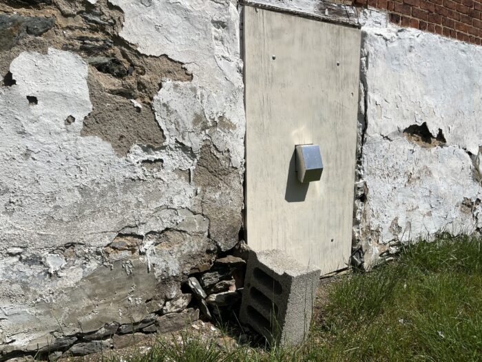 The damaged exterior of a building. A white, plaster-like substance chips off the wall of a basement area, with stone slabs coming dislodged toward the ground.