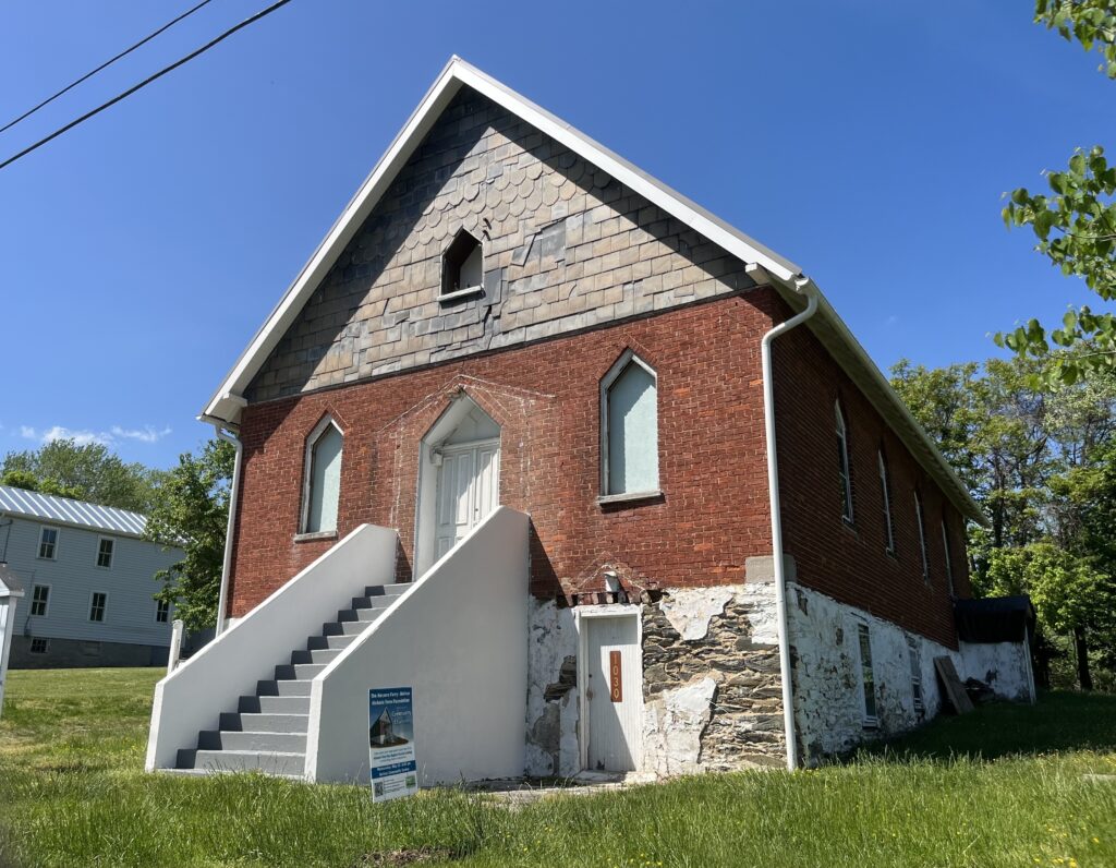 A small building with a faded facade and chipped roofing. It is brick, with a white staircase and sealed-off windows.