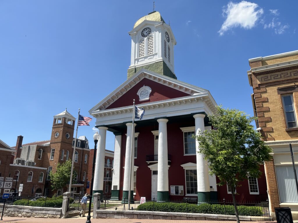 The American and West Virginia flags wave in front of the Jefferson County Courthouse. It stands tall, with four columns supporting a triangular top section and small steeple. A man walks toward the building from the sidewalk.