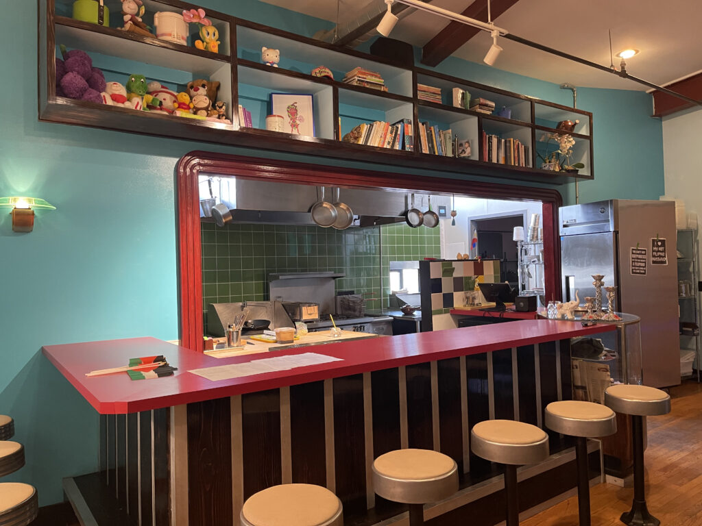 A bright and colorful diner counter with stools can be seen. Above the diner counter are shelves with an assortment of nick-knacks. Through the diner counter window is a kitchen.