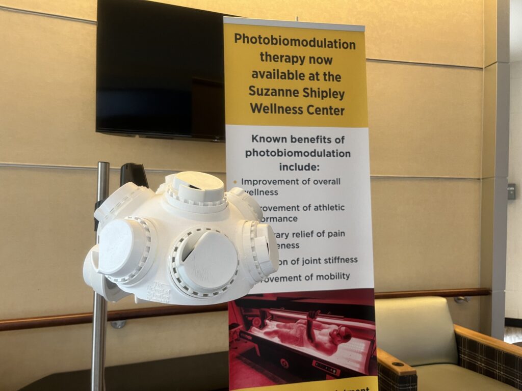 A helmet with many circular protrusions is attached to a pole at approximately head height if an individual wearing it was sitting. Behind it, a sign reads "Photobiomodulation therapy now available at the Suzanne Shipley Wellness Center."