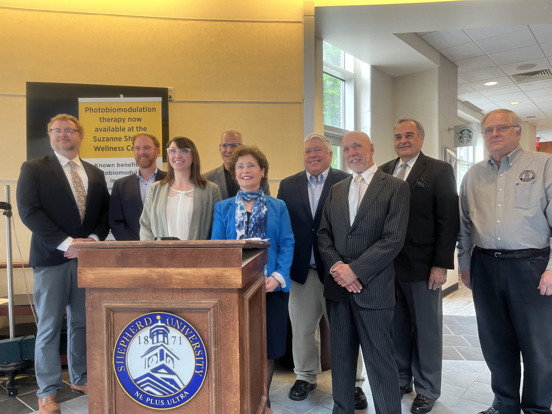 A group of people in suits stands behind a podium, smiling and posing for a picture. There are nine people split into two rows. On the podium the seal of Shepherd University is displayed prominently.