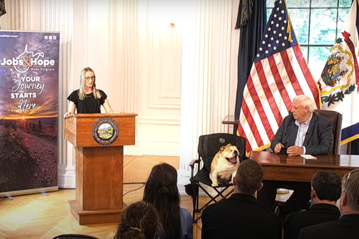 A woman stands at a podium, speaking into a microphone. Behind her, a sign reads "Jobs & Hope." To her side sits Gov. Jim Justice at his desk, with the American and West Virginia flags behind him. His pet bull dog Baby Dog is seated in a chair beside him, yawning. In front of Justice and the speaker are rows of people sitting down, visible only from the back.