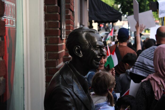 A bronze statue of actor Don Knotts sits in front of a red brick building. Behind Knotts' head can be seen two small Palestinian flags held by a smiling woman wearing a hijab. A crowd stretches on behind her, and another Palestinian flag can be seen in the background.