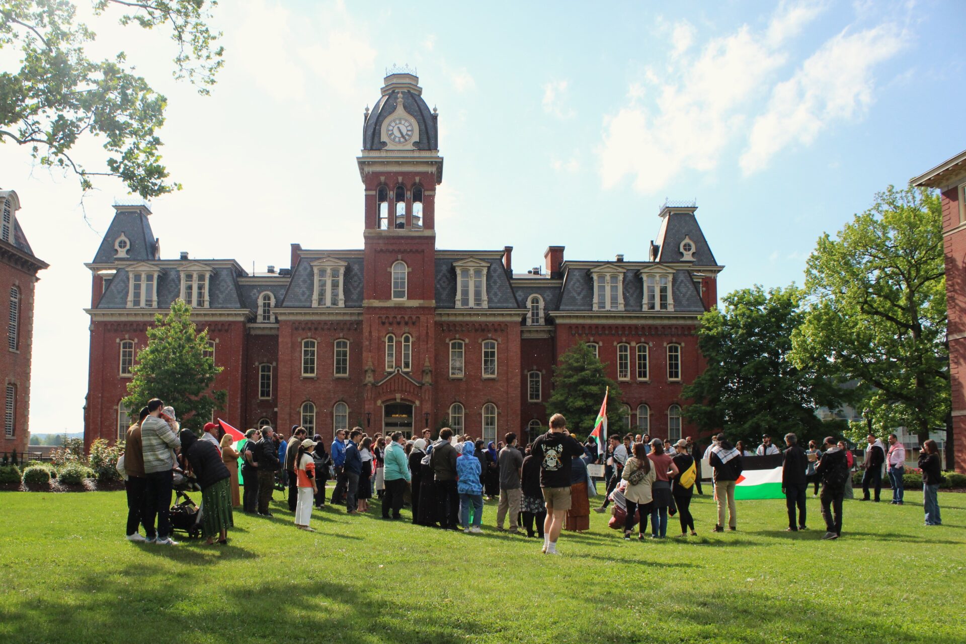 A group congregates on the lawn in front of a three story building with a clock tower at its center. It is a bright, sunny day with a blue sky and wispy white clouds. At the far right of the crowd two people hold a Palestinian flag. Closer to the foreground and apart from the crowd stands a family around a stroller.