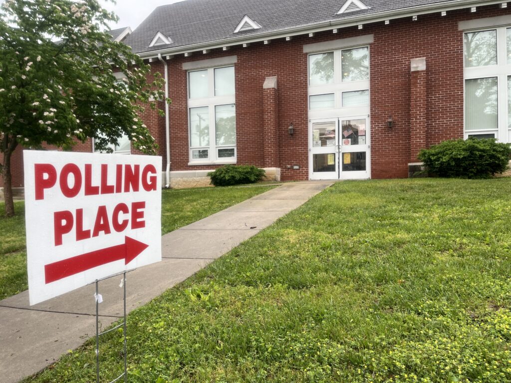 A sign reads "polling place" with a large arrow pointing to the front doors of a church. A sidewalk leads to the church doors, which are lined with election information documents and two small American flags.