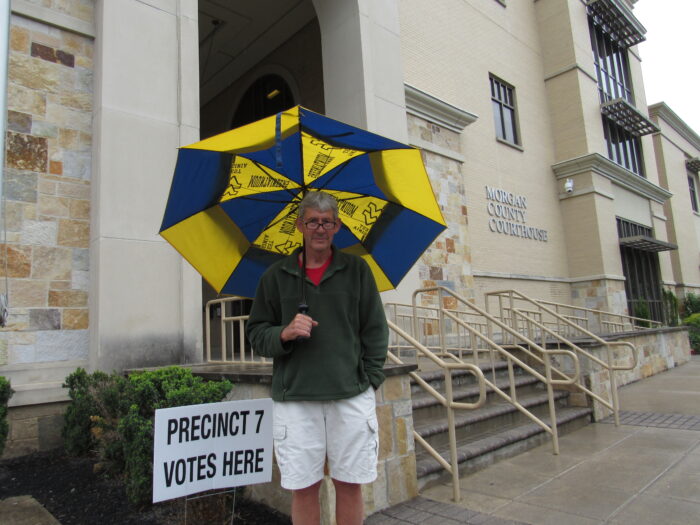 A man stands beneath an umbrella detailed with the logo of West Virginia University. Behind him is a sign that reads "Precinct 7 Votes Here" and the Morgan County Courthouse, with a stone staircase leading to a large archway.