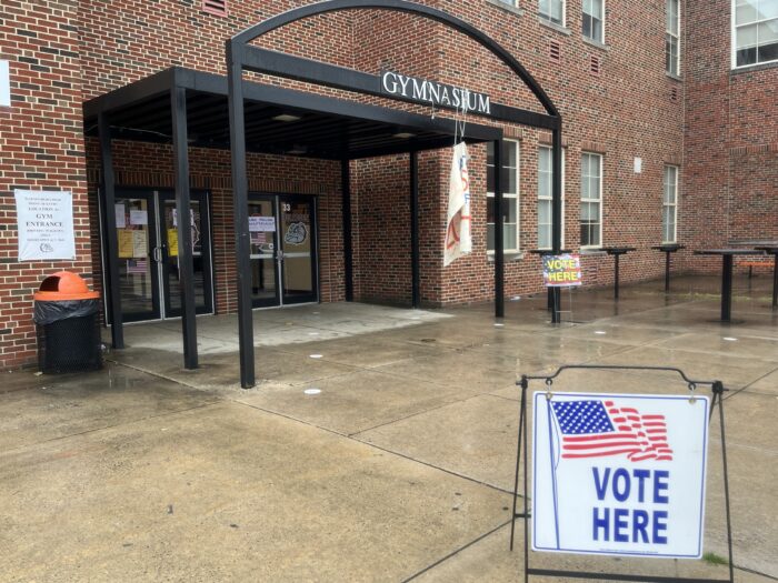 A sign with the American flag depicted on it reads "Vote Here." Behind it is an entryway labeled "Gymnasium." The sidewalk leading to the entryway is visibly wet from the rain.