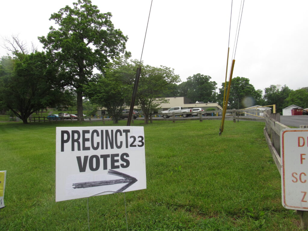 A sign reads "Precinct 23 Votes," with an arrow pointing toward a school parking lot. The school is slightly visible in the background.