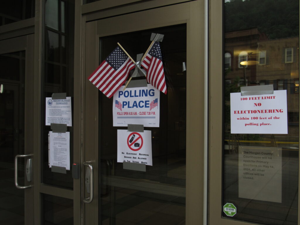 A dark-tinted glass door displays signs containing election information, including one sign that reads "Polling Place" and displays the polling location's hours of operation. Two miniature American flags are displayed prominently above the signs.