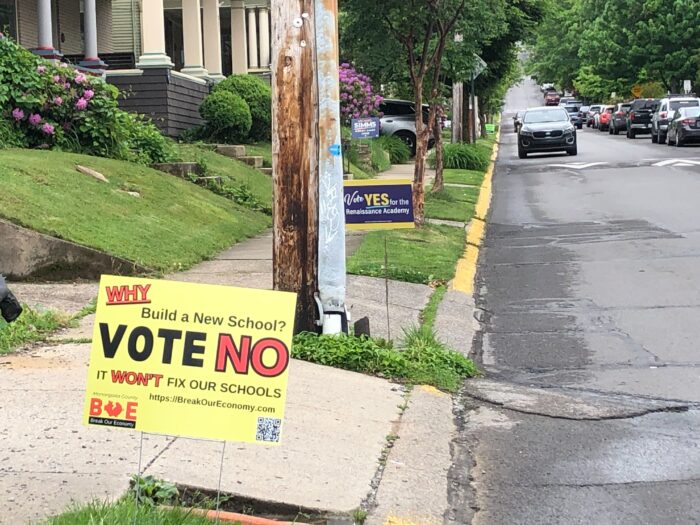 Two lawn signs with opposing positions on a school issue are placed on the same road. To the right of frame, a car is approaching frame. The sign closest to the camera has red and black text on a yellow background that reads "Why Build a New School? Vote NO IT WONT'T FIX OUR SCHOOLS" The sign further from the camera and partially obstructed by a power pole has white text on a blue background with a yellow YES that reads "Vote YES for the Renaissance Academy."