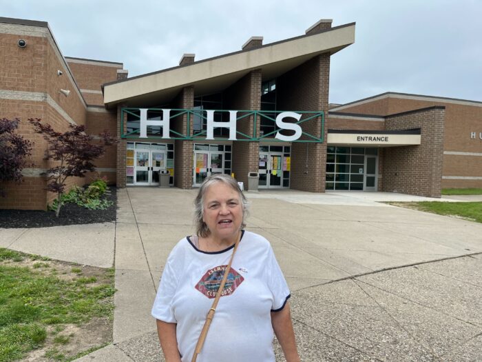 An older woman stands in front of Huntington High School in on May 14, 2024. She wears a white shirt and a purse hangs off her shoulder.