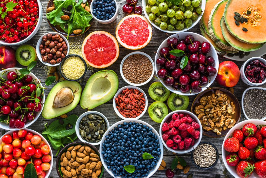 A colorful array of fruits and vegetables is displayed on a table. A large bowl of blueberries is visible at the center bottom of frame, with a bowl of raspberries to the right. There are cut kiwis, a bowl of cherries, an avocado and a grapefruit cut in half. There are a few bowls of nuts as well.