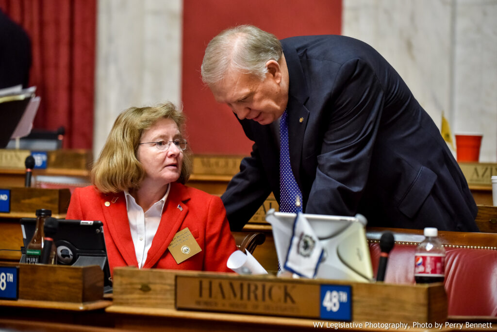 A woman in a red jacket and white collared shirt sits at a wooden desk as a man in a dark blue suit crouches down to speak with her.