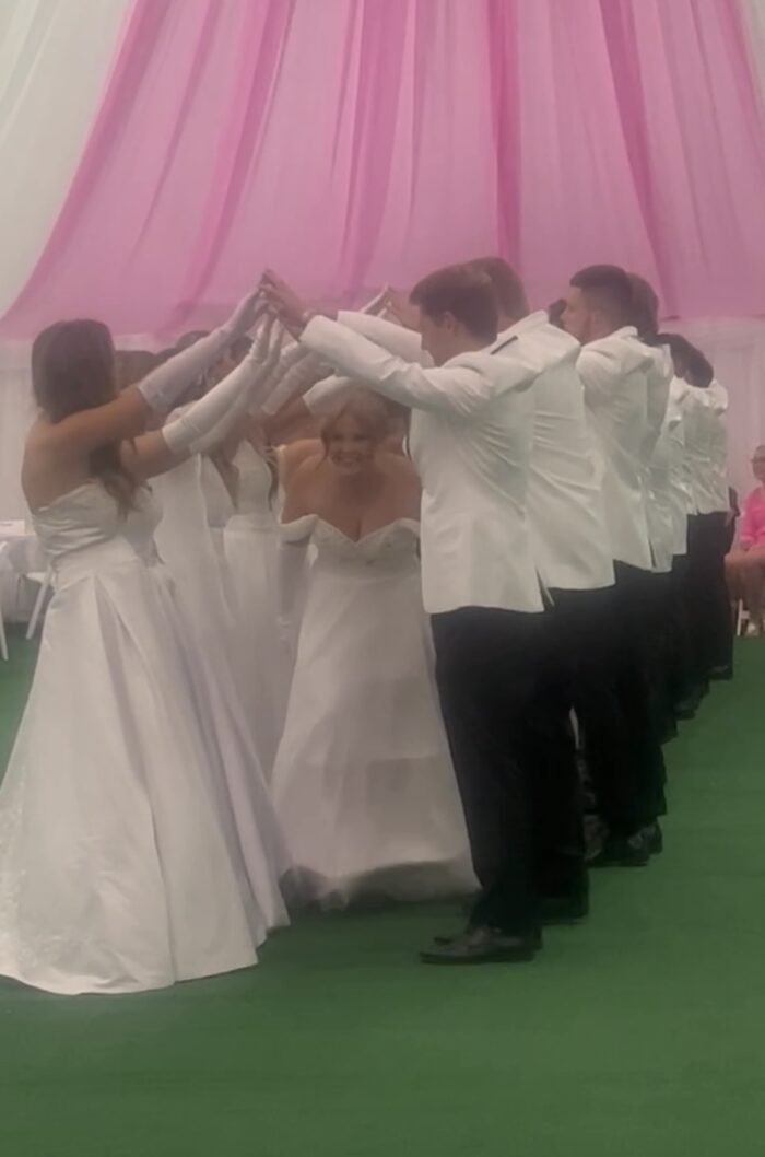 A close up shot of young people doing a formal dance. The group have their hands raised in a bridge style. The women wear white gowns, and the men wear white suit jackets and black slacks and shoes.