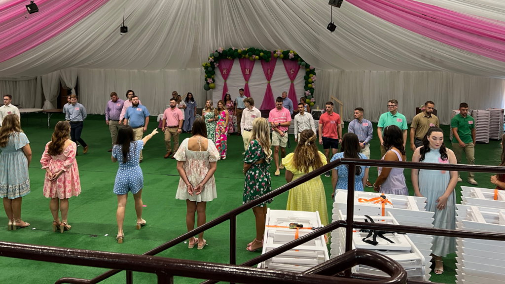More than two dozen young people stand on a floor covered in green carpet. Above them is a canopy of pink and white fabrics. All of the individuals are dressed formally in colorful, spring dresses or button up shirts and khaki shorts or pants.