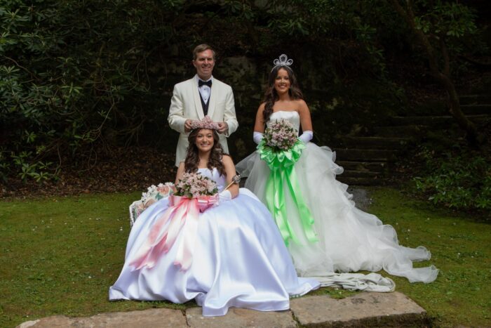 Three people smile for the camera. One man and two women. They are all dressed formally. The women are in white ball gowns, while the man wears a white suit with a black bow tie. One woman is seated, and a crown is being placed on her head by the man.