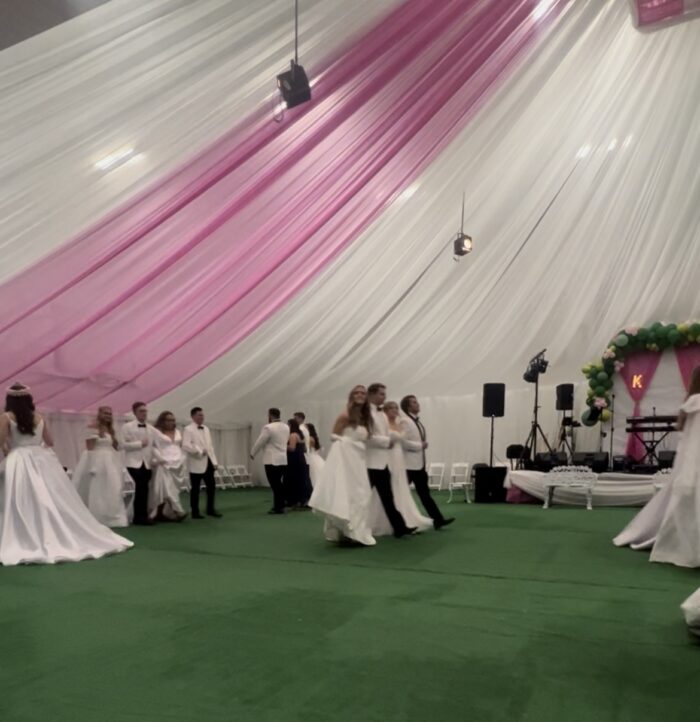A wide shot of individuals performing in a formal dance. All are dressed formally, the women in white gowns, and the men in white suits, black slacks, and black bow ties.