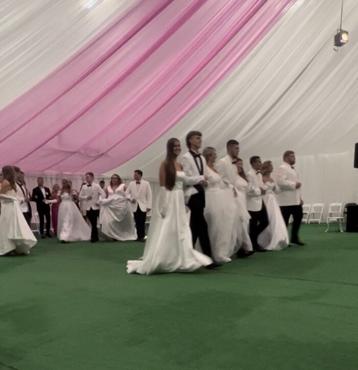 A closer shot of individuals performing in a formal dance. All are dressed formally, the women in white gowns, and the men in white suits, black slacks, and black bow ties.