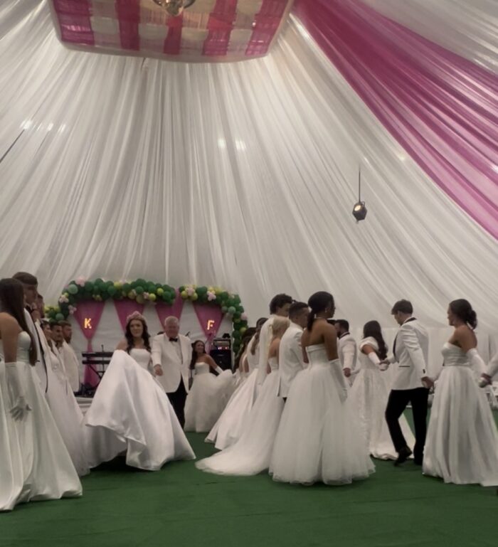Young people in formal dress dance together. The women are in white gowns, while the men wear white suit jackets and black slacks and shoes.