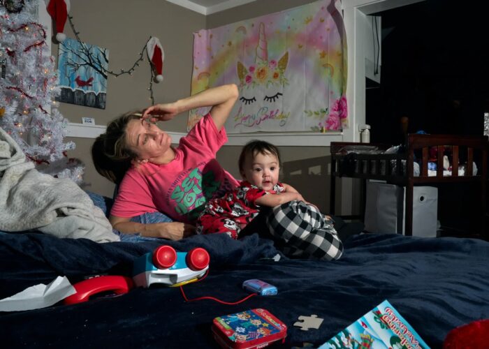 A woman leans on a bed as a small child sits next to her. The child looks toward the camera, while the woman looks off frame and lifts a hand to her head.