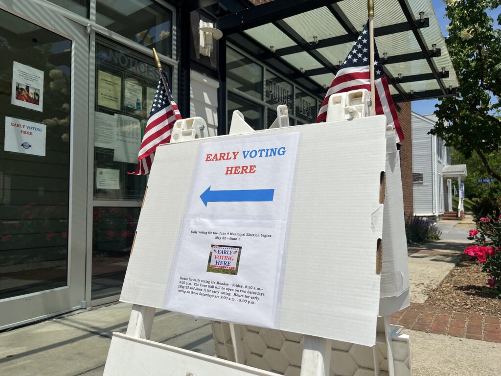 A sign bearing two miniature American flags reads "Early Voting Here," with an arrow pointing to the left. Beneath this is text explaining the days and hours of early voting for a municipal election.