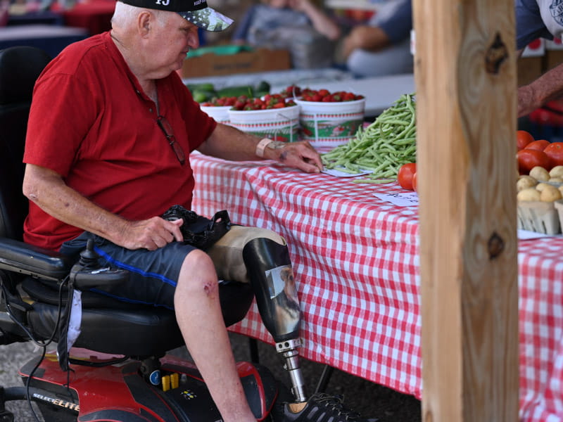 An elderly man in a wheelchair and with a prosthetic leg looks at food displayed on a table.