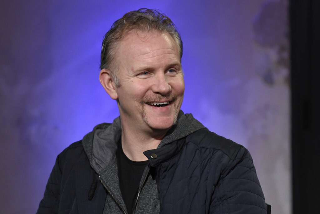 A middle age man smiles in a photograph. He looks off frame as if in an interview. He wears a black jacket.