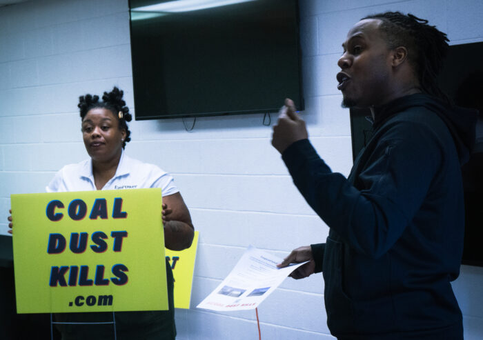 Two people, one woman and one man, speak before an unseen crown in a room. The woman is holding a yellow sign that reads, "Coal Dust Kills dot com." The man next to her is speaking passionately and holding a white paper.