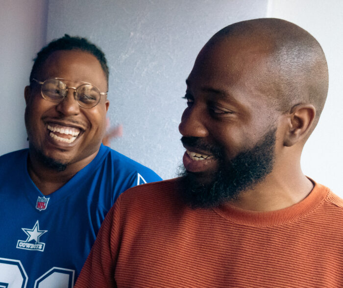 Two men laugh in a photograph. The photo is a close up shot. One man wears glasses and a blue shirt. The other man is balding, has a beard, and wears an orange shirt.