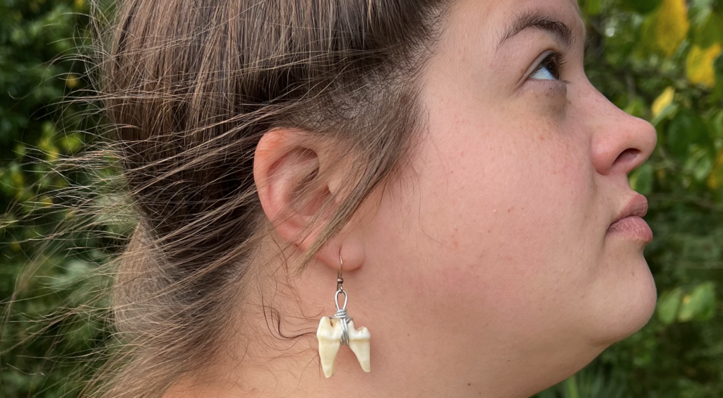 A close-up, profile image of an adult woman with brown hair. She looks up off camera, and the focus of the photo is on her earrings. Her earrings are teeth wrapped around metal wire.