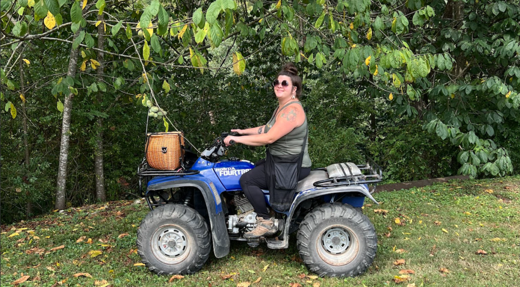 An adult woman sits on a four-wheeler wearing sunglasses. She smiles for the camera. She wears a tank top shirt and black pants. She sports a messy bun of hair. There is a wicker basket on the front of the four-wheeler.