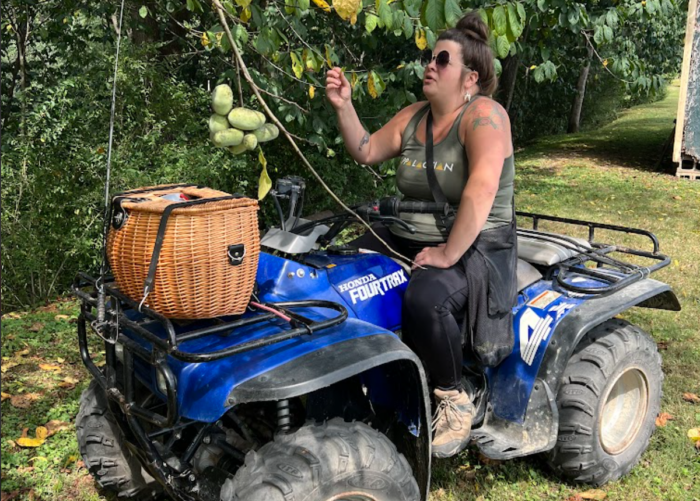 An adult woman sits on a four-wheeler wearing sunglasses. She smiles for the camera. She wears a tank top shirt and black pants. She sports a messy bun of hair. There is a wicker basket on the front of the four-wheeler. She also picks pawpaws from a tree.