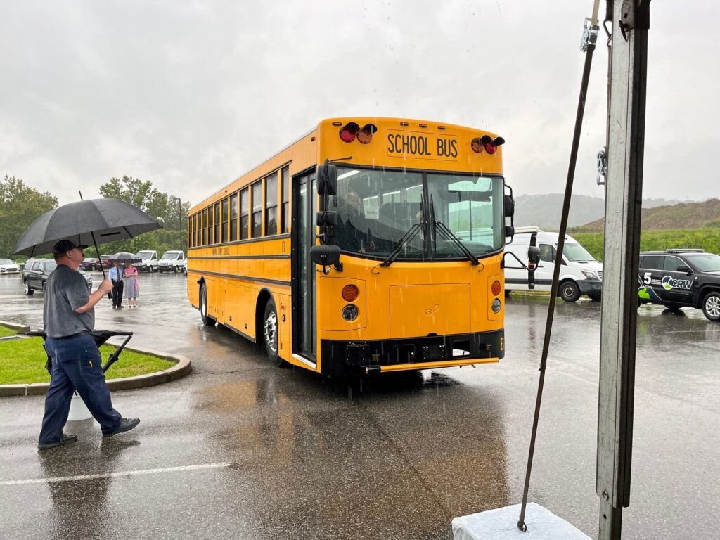 A yellow, electric school bus is shown parked on the side of the road. A man holding an umbrella approaches it. The sky is overcast, and it is raining.