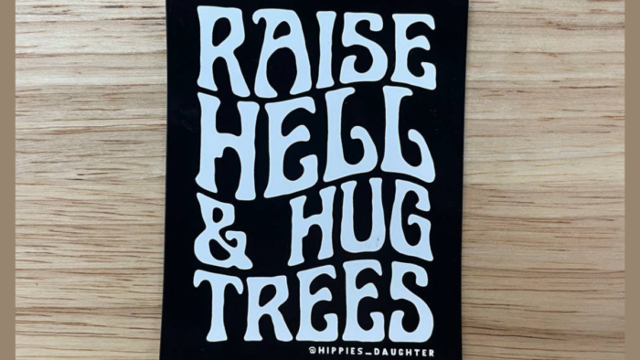 A sticker is shown on a wooden table. The sticker is black with white letters featured on it. The letters read, "Raise Hell and Hug Trees." Underneath the text is smaller text that reads, @HIPPIES_DAUGHTER.