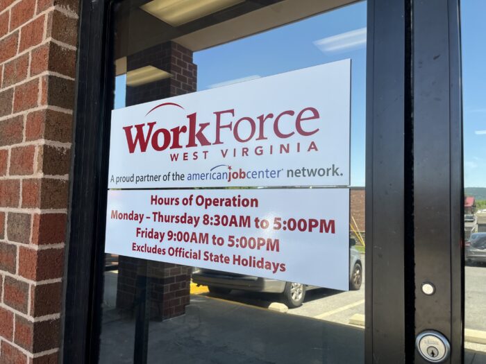A sign on a door reads "WorkForce West Virginia," with its hours of operation listed below. The door is placed within a brick building facade.