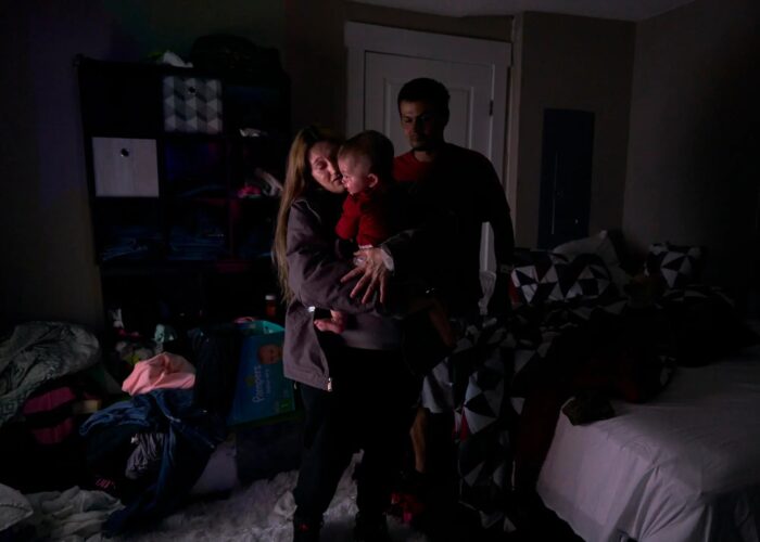 A woman holds a small child as a man stands behind her. The photograph is dark, as if there wasn't much light in the room.